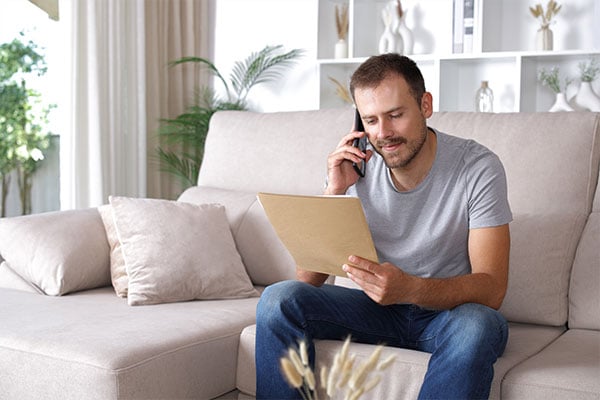 A man sitting on a couch talking on the phone while reviewing insurance documents.